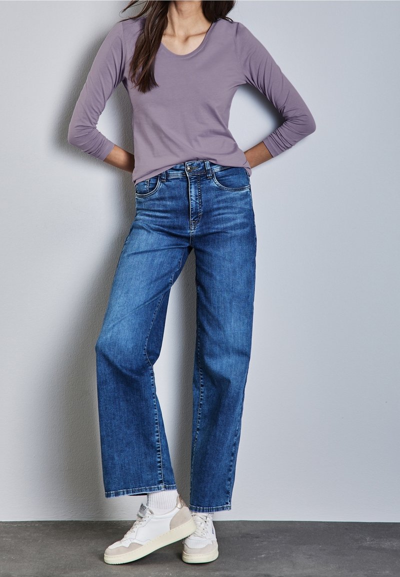 Lila långärmad skjorta matchad med högmidjade, raka blå jeans. Skorna har vita och beige detaljer i en sneakersdesign.