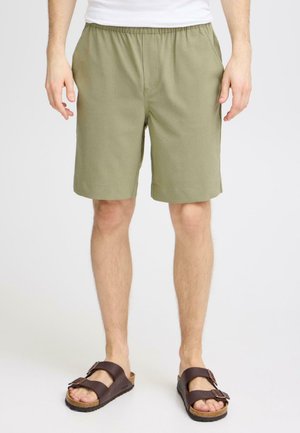 Mann iført olivengrønne shorts med elastisk midje og brune sandaler med dobbel stropp, står foran en ensfarget bakgrunn.