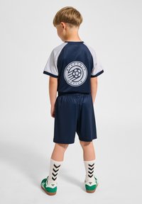 Marinblå och vit sportoutfit med logotryck på ryggen, bestående av shorts och en kortärmad topp, kombinerad med mönstrade strumpor och gröna skor.