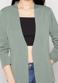 Ljusgrön kavaj med sjalkrage och rynkade ärmar, bärs över en svart åtsittande topp. Har en slät, texturerad tyg.