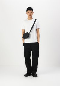 Sac en bandoulière noir en cuir, porté avec un t-shirt blanc et un pantalon cargo noir. Les chaussures sont noires et épaisses avec une semelle plate.