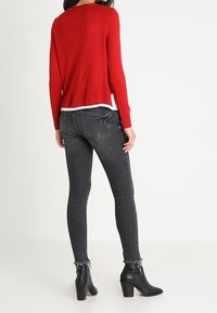 Pull en maille rouge à manches longues, coupe décontractée, superposé sur un haut blanc. Jeans skinny noirs taille haute avec des ourlets effilochés et des bottines noires.