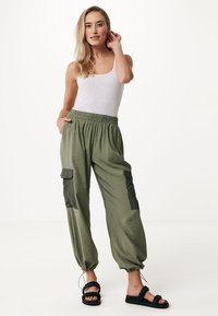 Pantaloni leggeri verde oliva con cintura elastica, tasche laterali e caviglie affusolate, abbinati a una canotta bianca e sandali neri.