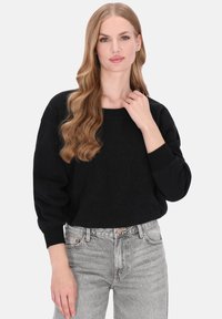 Zwarte oversized trui met gestructureerd patroon, brede halslijn en geribde mouwen, gecombineerd met lichtgrijze high-waisted jeans.