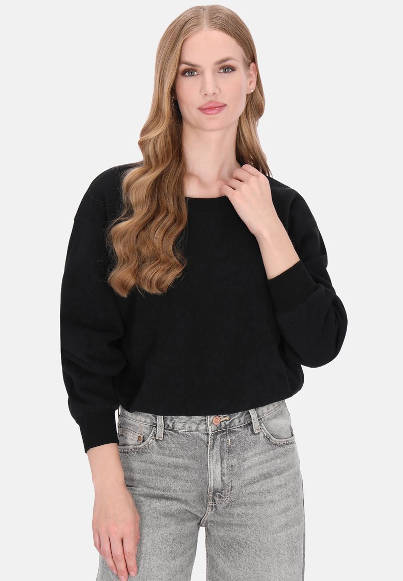 Zwarte oversized trui met gestructureerd patroon, brede halslijn en geribde mouwen, gecombineerd met lichtgrijze high-waisted jeans.