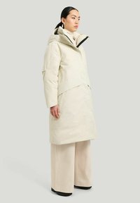 Beige vattentät parka med en stor huva, framfickor och en strukturerad silhuett. Bärs med ljusfärgade vida byxor och svarta skor.