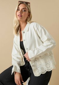 Vit denimjacka med dekorativ spetskant, silverfärgade knappar, långa ärmar och en avslappnad passform. Svart topp och byxor fulländar outfiten.