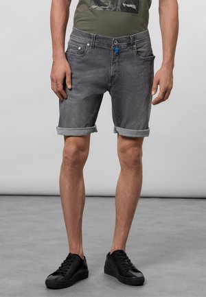 Homme portant un short en denim gris foncé retroussé avec un détail de bouton bleu, des baskets noires et un t-shirt graphique vert olive.