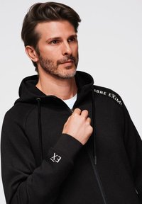 Sudadera con cremallera negra y capucha con cordón, con un logotipo blanco en el hombro izquierdo y en el puño. Tejido suave y texturizado, ajuste relajado.