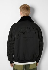 Svart bomberjacka med fuskpälskrage, quiltad textur och präglad design på baksidan. Ribbad mudd och midja.