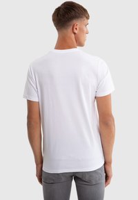 Giovane uomo con capelli castani corti, indossa una semplice T-shirt bianca a maniche corte e jeans grigi, visto di spalle su uno sfondo grigio.