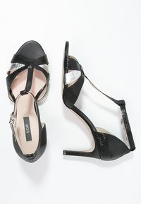 Black heeled sandals with a textured finish, metallic accents, and crisscross straps. Features a sleek design and a narrow heel.