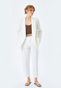 Witte linnen blazer over een bruine tanktop, witte broek met een geweven riem, en lichte sandalen met gouden accenten. Soepele textuur en een getailleerde pasvorm.