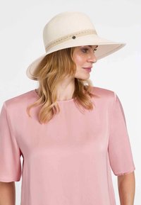 Sombrero de ala ancha en color crema con una superficie texturizada y una banda trenzada. El sombrero cuenta con un botón decorativo y se combina con una blusa rosa.