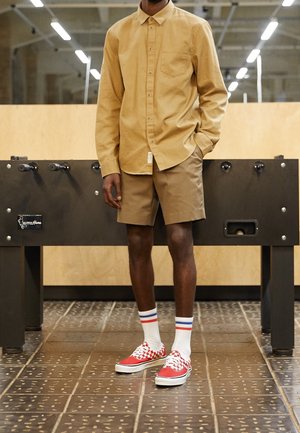 Tan dlouhý rukáv, hnědé šortky, červené kostkované tenisky a bílé ponožky s modrými a červenými pruhy. Stojí vedle stolního fotbálku.