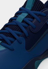 Μπλε αθλητικό παπούτσι με σταυρωτά κορδόνια, δίχτυ στη γλώσσα και επενδύσεις από δέρμα σε διάφορες αποχρώσεις του μπλε, εμφανιζόμενο σε λεπτομερή κοντινή λήψη.