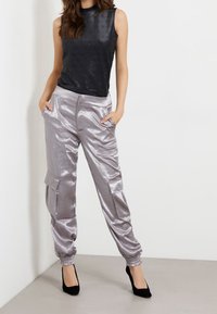 Mujer vestida con una blusa negra sin mangas con un diseño, pantalones cargo plateados brillantes con puños elásticos y zapatos de tacón negros, de pie con las manos en los bolsillos.