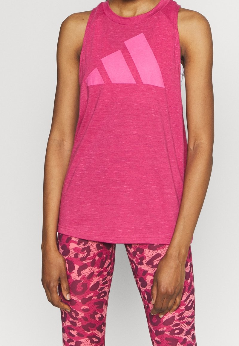 Personne portant un débardeur rose avec un grand logo et un legging rose à motifs, debout devant un fond clair uni.