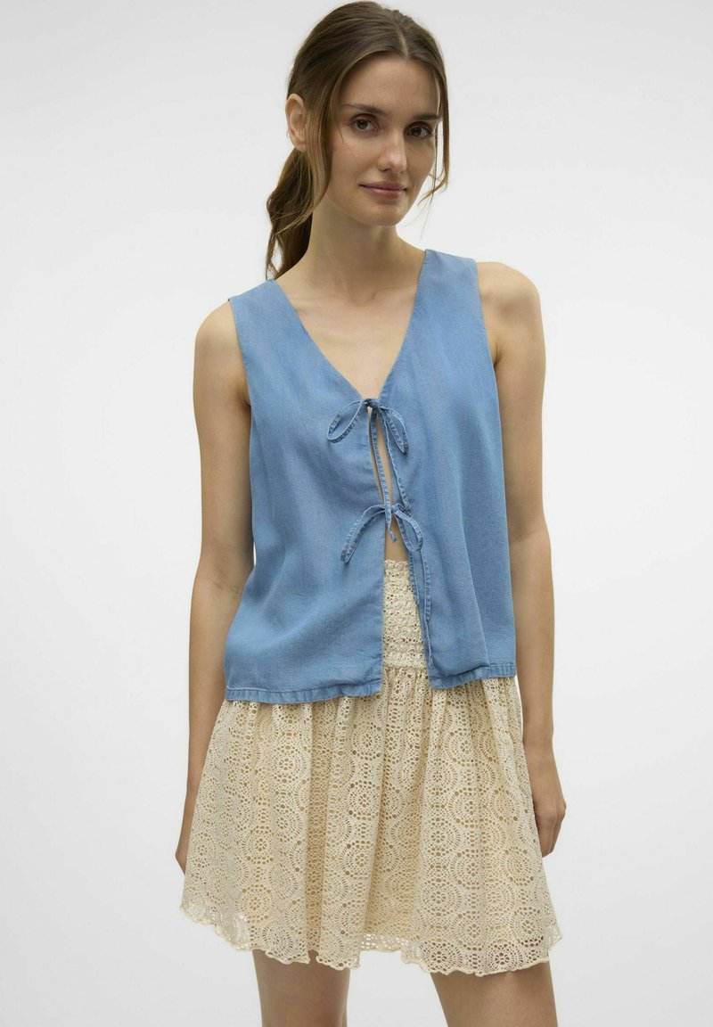 Ljusblå ärmlös topp med V-ringning och framsluten knytning, tillsammans med en beige spetskjol med ett cirkulärt mönster.