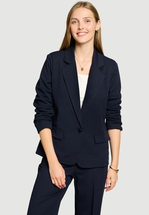 Jeune femme aux longs cheveux blonds portant un blazer bleu marine et un pantalon, souriant devant un fond uni gris clair.