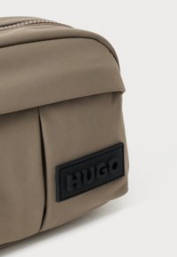 Brun nylon taske med en rund form, prydet med et sort gummilogopatch med "HUGO" og en lynlåslukning øverst. Glat tekstur.