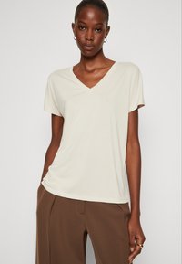 T-shirt beige chiaro con scollo a V, maniche corte, tessuto morbido, vestibilità leggermente ampia. Abbinato a pantaloni marroni a vita alta.