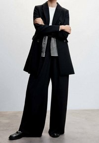 Zwarte double-breasted blazer over een wit-zwart gestreept shirt, gecombineerd met wijde zwarte broek en glanzende zwarte loafers.