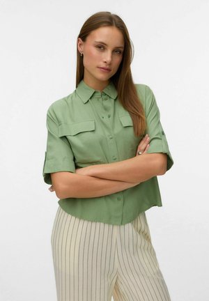 Ung kvinde med langt brunt hår iført en lysegrøn skjorte med knapper og ærmerne rullet op samt beige bukser med lodrette striber, armene krydsede.