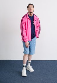 Pink zip-up jacket with black stripes, navy t-shirt, light blue shorts, white sneakers with black stripes, and white socks.