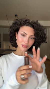 Make-up stick in een bruine tube, vastgehouden door een persoon met krullend haar, die een wit shirt met lange mouwen draagt. Korte, verzorgde nagels met witte tips.