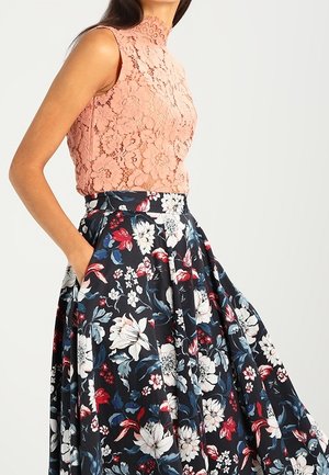 Kobieta ubrana w koralowy koronkowy top bez rękawów z wysokim kołnierzem oraz czarną spódnicę z wysokim stanem ozdobioną dużym, wielokolorowym kwiatowym wzorem.
