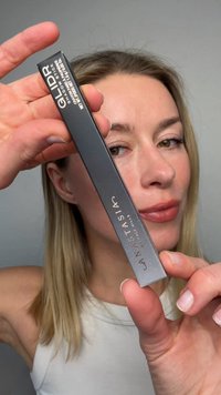 Frau mit blonden Haaren hält eine schwarze Anastasia Beverly Hills GLDR Eyeshadow Stick-Box nah an ihrem Gesicht vor einem grauen Hintergrund.