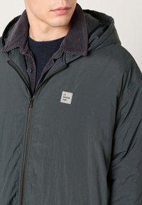 Muž v tmavosivej bunde na zips s kapucňou a golierom z kordurovej látky, pod ňou námornícka košeľa a malá nášivka s nápisom "LA MAISON AMV" na hrudi.