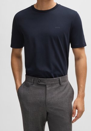 Mann trägt ein eng anliegendes, marineblaues BOSS-T-Shirt, das in hoch taillierte, graue, maßgeschneiderte Hosen mit Seitentaschen gesteckt ist.