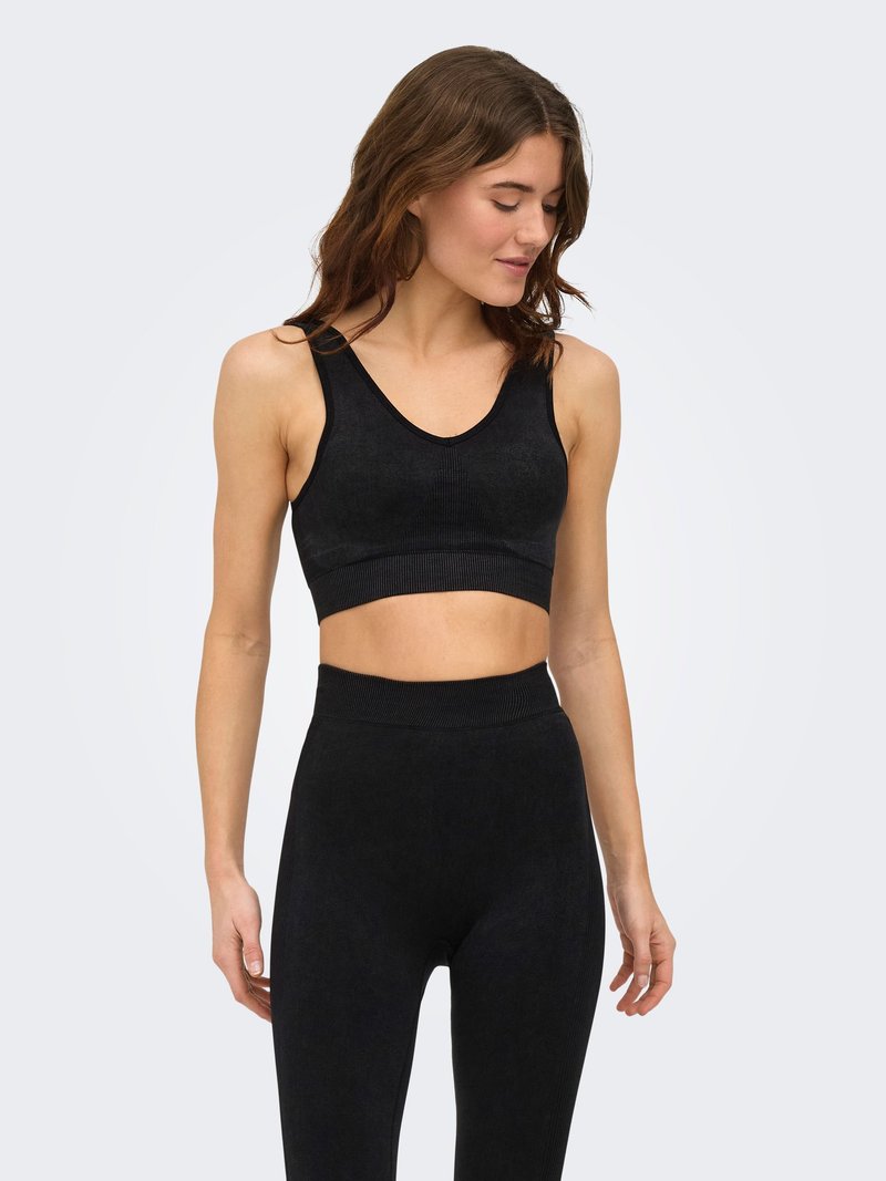 Femeie cu păr castaniu ondulat, purtând un top negru fără mâneci și colanți cu talie înaltă, privind în jos și zâmbind ușor, pe un fundal simplu.