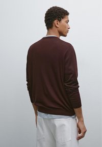 Maglione in bordeaux a maglia con scollo rotondo, maniche lunghe e texture morbida, indossato sopra una camicia grigia chiara e abbinato a pantaloni bianchi.