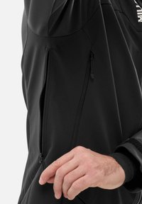 Hand adjusting zipper on black hooded jacket with zippered pockets and Velcro wrist strap on left sleeve.