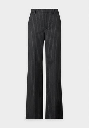 Femme portant un haut noir asymétrique à manches courtes avec un pantalon noir à jambes larges à rayures fines, debout avec une main dans la poche.