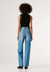 Top negro sin mangas con detalle de ojales en los costados, combinado con jeans de pierna ancha en azul claro con bolsillos traseros y una etiqueta de cuero roja.