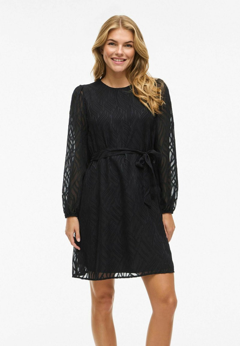 Black dress with long, sheer sleeves, featuring a textured leaf pattern and a belt at the waist. Knee-length silhouette with a round neckline.