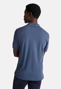 Muž s krátkými kadeřavými vlasy stojí zády, má na sobě modrou polo košili s krátkým rukávem a tmavé kalhoty na bílém pozadí.