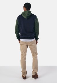 Gevoerd sweatshirt met een marineblauw torso en groene mouwen, met een capuchon met trekkoord, elastische zoom, en beige broek. Bruine schoenen zichtbaar.