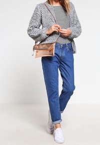 Kobieta w teksturowanej szarej kurtce, w paskowanej koszuli, niebieskich spodniach z mankietami, białych trampkach i niosąca metalicznie różową torebkę crossbody.