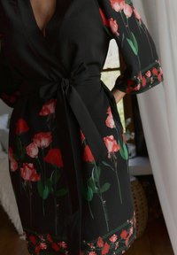 Zwarte bloemen wikkelrobe met rode en roze rozenpatronen, lange mouwen en een stoffen strik in de taille. Gevoed zwarte achtergrond.