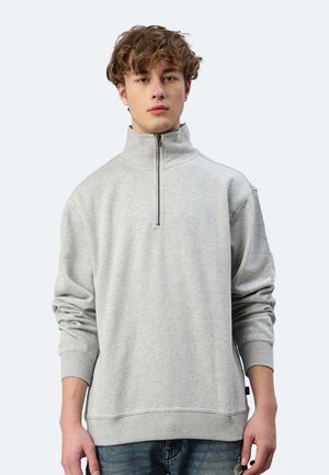 Junger Mann mit lockigem Haar, der ein hellgraues Sweatshirt mit Viertel-Reißverschluss und eine blaue Jeans trägt, vor einem schlichten hellen Hintergrund steht.