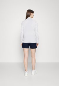 Biela blúzka s dlhým rukávom a textúrovanými rukávmi, skombinovaná s tmavomodrými šortkami. Model má na sebe biele športové topánky a je otočený chrbtom k fotoaparátu.