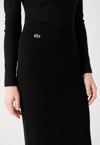 Personne portant une jupe côtelée noire avec un logo crocodile vert à la taille, associée à un haut noir à manches longues.
