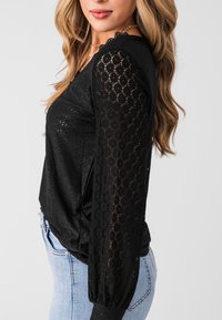 Schwarze Bluse mit langen, durchsichtigen Spitzenärmeln mit floralem Muster, einer taillierten Passform und glatter Textur, kombiniert mit hellblauen Jeans.
