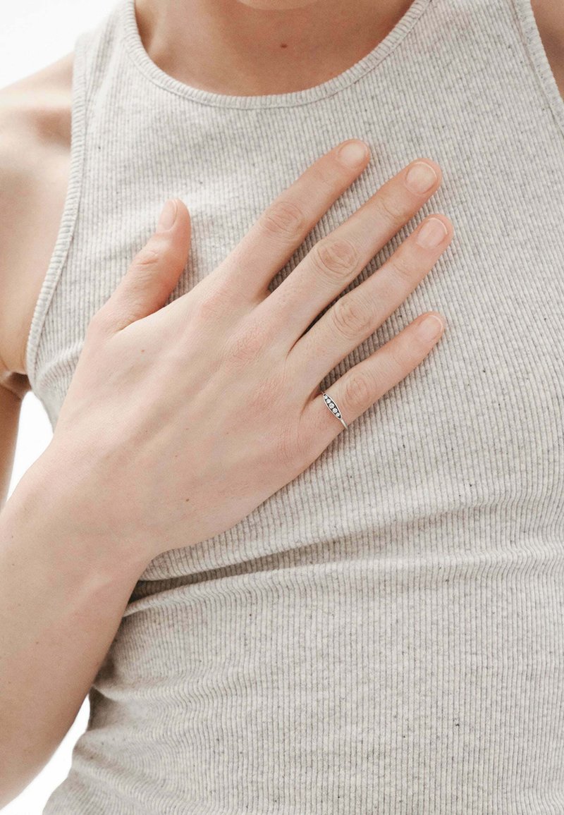 Anillo de plata con pequeñas piedras transparentes, colocado sobre una mano que descansa en una camiseta sin mangas de rayas en color gris claro. El tono de piel es claro. Textura suave en general.
