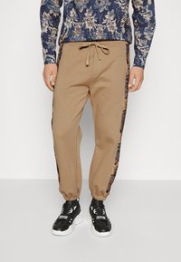 Bruna sweatpants med elastisk midja och mudd, med blommönstrade sidodetaljer i mörka färger. Matchas med svarta sneakers.